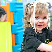 Isabella is registered to the contest to win money with this photo: background, black_shirt, blonde_hair, blue, casual_clothing, child, colorful, fun, happy, indoor, orange, people, play, playground, ribbons, slide, smiling, toddler, yellow, young_children