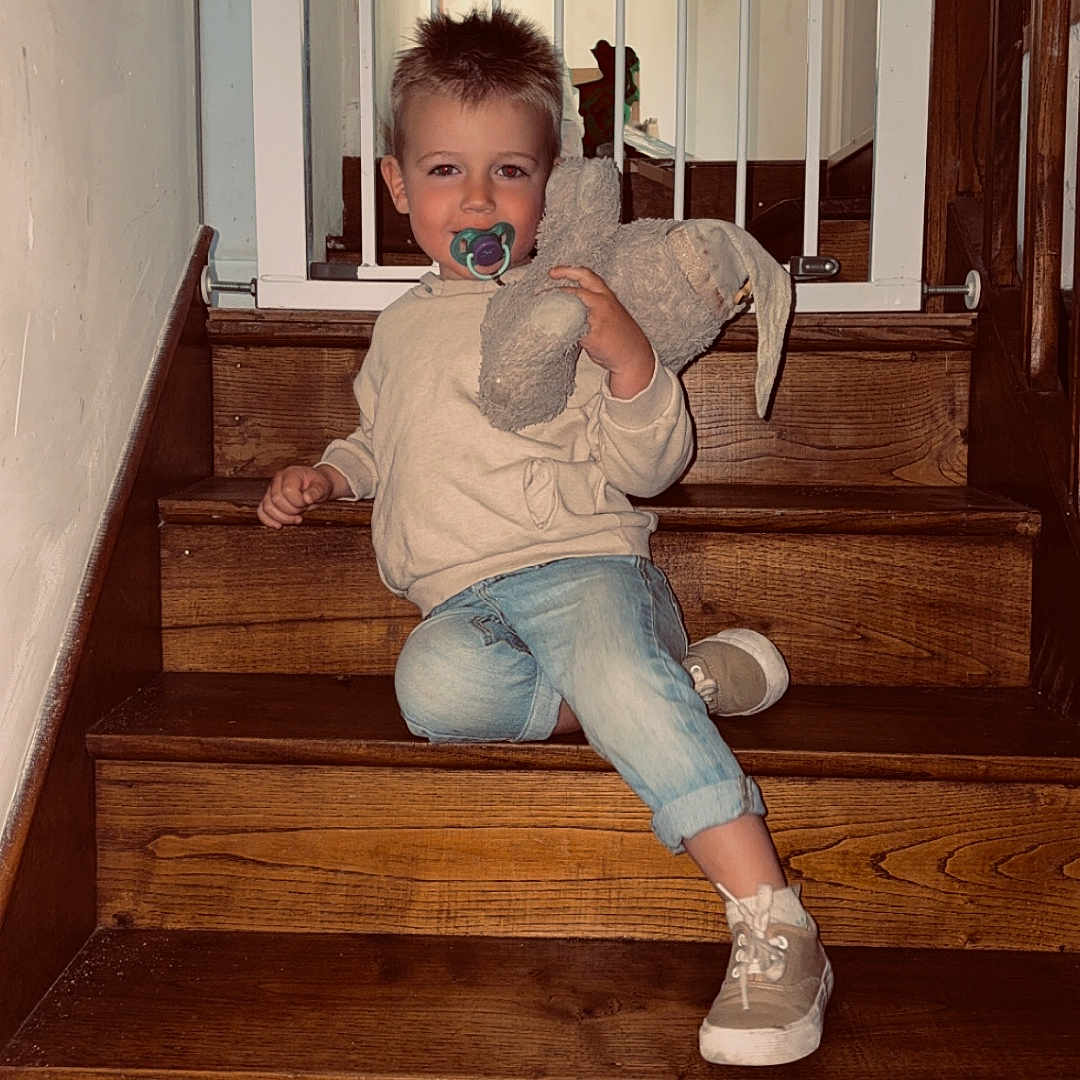 Tiago participe au concours pour gagner de l'argent avec cette photo : baby_gate, casual_clothing, child, comfort, cozy, cute, home, indoor, pacifier, person, plush_toy, portrait, seated, smiling, sneakers, stairs, toy, wood, wooden_stairs, young_child