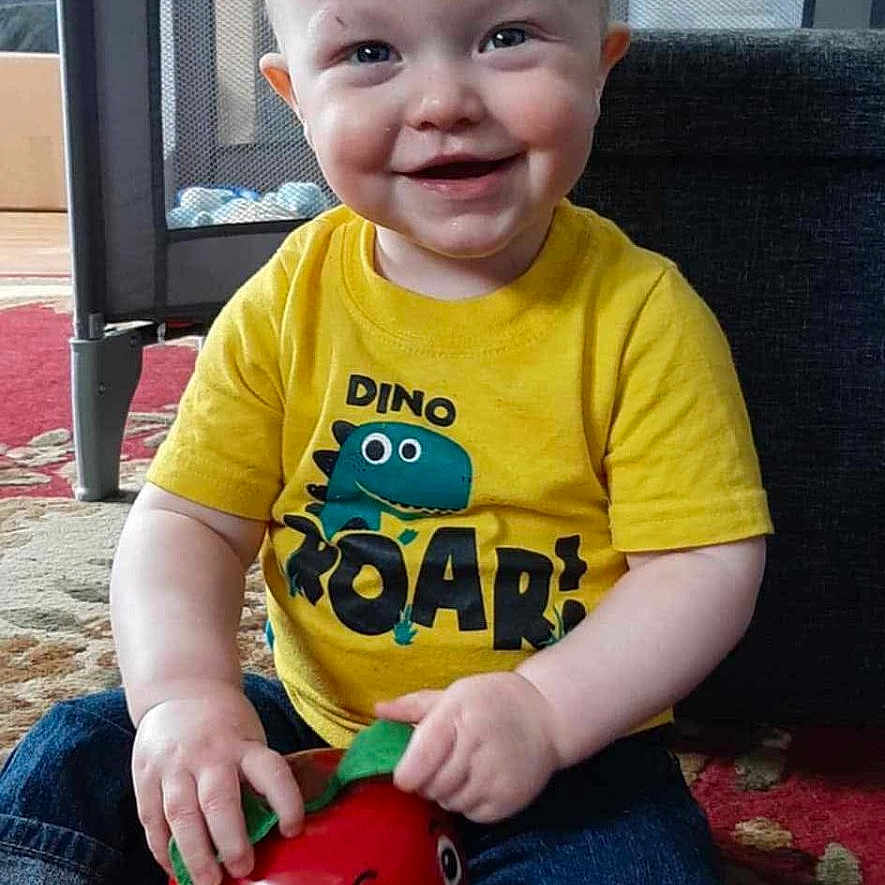 Nicholas is registered to the contest to win money with this photo: carpet, child, cute, dinosaur_graphic, furniture, happy, home, indoor, jeans, person, playpen, portrait, red_apple, route_66_sign, sitting, smiling, socks, toddler, toy, yellow_tshirt