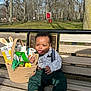 Jalen is registered to the contest to win money with this photo: baby, child, bench, outdoor, park, basket, snacks, toy, grass, trees, sunlight, clothing, playful, expression, tongue, person, green_pants, brown_shoes, curly_hair, wood