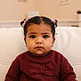 bow, child, couch, cozy, cute, earrings, expression, face, girl, hair_ties, home_interior, indoor, maroon_sweater, pigtails, portrait, seated, toddler, warm_lighting, white_couch, young_child