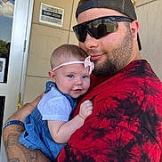 Bonnie joined the competition — help win amazing prizes! baby, child, man, holding, outdoor, building, door, headband, smile, tattoo, sunglasses, red_shirt, blue_dress, arm, person, portrait, casual, happy, closeup, daylight