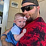 baby, child, man, holding, outdoor, building, door, headband, smile, tattoo, sunglasses, red_shirt, blue_dress, arm, person, portrait, casual, happy, closeup, daylight