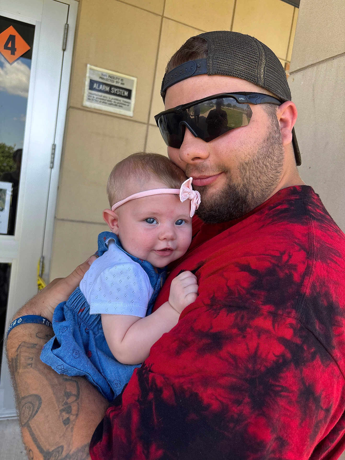 Bonnie joined the competition — help win amazing prizes! baby, child, man, holding, outdoor, building, door, headband, smile, tattoo, sunglasses, red_shirt, blue_dress, arm, person, portrait, casual, happy, closeup, daylight