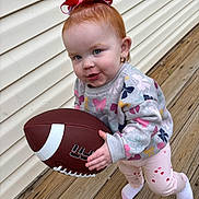Elsie joined the competition — help win amazing prizes! toddler, child, football, red_hair, bow, sweater, pink_pants, socks, outdoor, wooden_deck, wall, playful, cute, portrait, baby, person, holding, sport, standing, fun