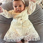 Camryn is registered to the contest to win money with this photo: baby, infant, white_dress, headband, lace, sitting, cushion, gray, soft, cute, raising_hand, footwear, shoes, indoors, portrait, child, smiling, elegant, clothing, person