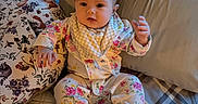 Camryn is registered to the contest to win money with this photo: baby, infant, pajamas, floral_pattern, bed, pillow, plaid_bedspread, blanket, cute, indoors, child, person, sleepwear, soft_lighting, cozy, young_child, portrait, sitting, home, curious