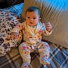 baby, infant, pajamas, floral_pattern, bed, pillow, plaid_bedspread, blanket, cute, indoors, child, person, sleepwear, soft_lighting, cozy, young_child, portrait, sitting, home, curious