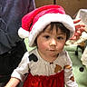 baby, child, toddler, santa_hat, christmas, holiday, portrait, indoor, smiling, facial_expression, dark_hair, brown_eyes, red_dress, festive_clothing, adult, hand, furniture, home, decorations, photography