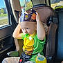 child, car_seat, pacifier, burger_king_crown, green_shirt, sippy_cup, car_interior, sunlight, seatbelt, toddler, window, yellow, blue, plastic, sitting, casual_clothing, daylight, young_child, portrait, curious