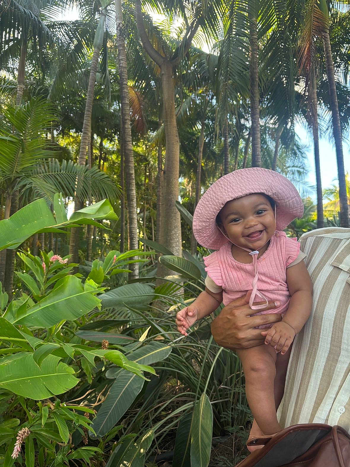 Kalycia a rejoint le concours — aidez-le/la à gagner de superbes lots ! baby, child, pink_hat, pink_clothing, smiling, happy, person, adult_hand, striped_shirt, tropical, greenery, plants, leaves, palms, outdoor, sunlight, nature, portrait, infant, holding