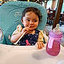 baby, child, clothing, cute, face, feeding, hand, high_chair, indoor, lighting, playful, polka_dot, restaurant, seat, sippy_cup, smile, straw, table, toddler, wooden_furniture