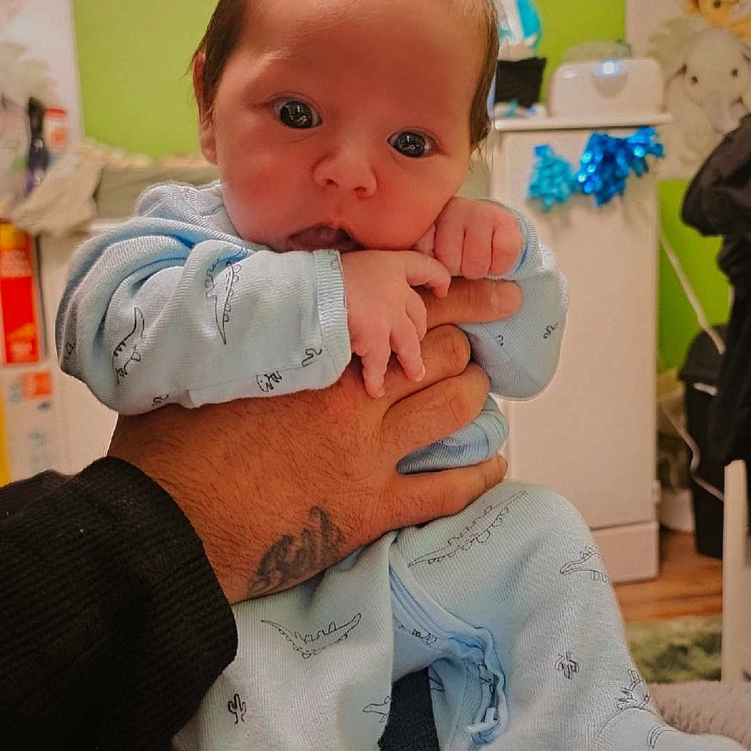 Ezekiel is registered to the contest to win money with this photo: baby, infant, child, portrait, indoors, nursery, held, hands, smiling, calm, blue_outfit, onesie, baby_clothing, animal_pattern, green_wall, stuffed_animals, home, family, closeup, photography