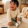 baby, child, smiling, sitting, indoor, carpet, plush_toy, teddy_bear, plant, woven_basket, window, natural_light, clothing, long_sleeve, pants, sock, face, person, home_decor, cute
