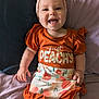 baby, child, smiling, headband, orange_clothing, sitting, happy, portrait, infant, bed, pillow, cute, human, apparel, indoors, person, face, skin, expression, comfort