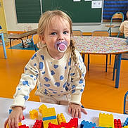 Kamilia participe au concours pour gagner de l'argent avec cette photo : toddler, child, pacifier, building_blocks, table, classroom, toys, play, blonde_hair, braid, heart_pattern, sweater, colorful, indoor, education, learning, chair, clock, blackboard, floor
