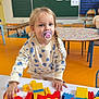 toddler, child, pacifier, building_blocks, table, classroom, toys, play, blonde_hair, braid, heart_pattern, sweater, colorful, indoor, education, learning, chair, clock, blackboard, floor