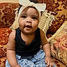baby, child, headband, bow, black_top, denim_pants, sofa, cushion, patterned_fabric, indoor, portrait, smiling, seated, curly_hair, cute, infant, home, cozy, person, toddler
