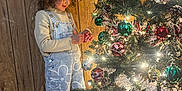 Estrella is registered to the contest to win money with this photo: child, christmas_tree, curly_hair, decorating, festive, flower_pattern, green, holiday, indoor, lights, ornaments, overalls, pine_cones, plaid, red, silver, smiling, socks, tree_skirt, wooden_wall