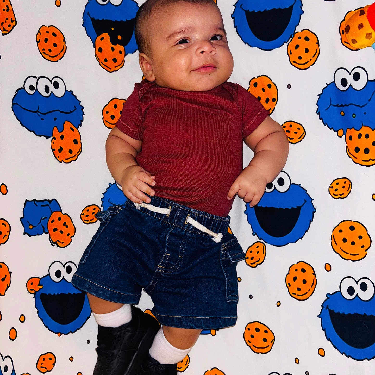 Eleggúasdiel joined the competition — help win amazing prizes! baby, black_shoes, blanket, casual_clothing, child, colorful, cookie_monster, cookie_pattern, cute, denim_shorts, happy, indoors, infant, lying_down, maroon_shirt, patterned_background, playful, portrait, smiling, white_socks