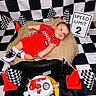 baby, birthday, black_and_white, cake, celebration, checkered_flag, decorations, footwear, indoor, infant, orange_cone, person, plush_cushion, portrait, racing_theme, red_clothing, smiling, speed_limit_sign, tire, toy