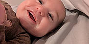 Oliver is registered to the contest to win money with this photo: baby, smiling, face, person, bed, pillow, brown_clothing, lying_down, indoors, happy, child, infant, closeup, skin, head, eyes, mouth, cheeks, sleepwear, blanket