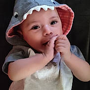 Ivan joined the competition — help win amazing prizes! baby, child, hood, shark_costume, cute, infant, face, hands, eyes, clothing, smile, portrait, lying_down, dark_background, costume, cute_outfit, baby_garment, adorable, baby_boy, baby_girl