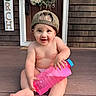 Alexander is registered to the contest to win money with this photo: baby, child, smiling, cap, water_bottle, porch, wooden_floor, wreath, door, sign, happy, sitting, outdoor, cute, infant, bare_chest, playful, person, holding, summer