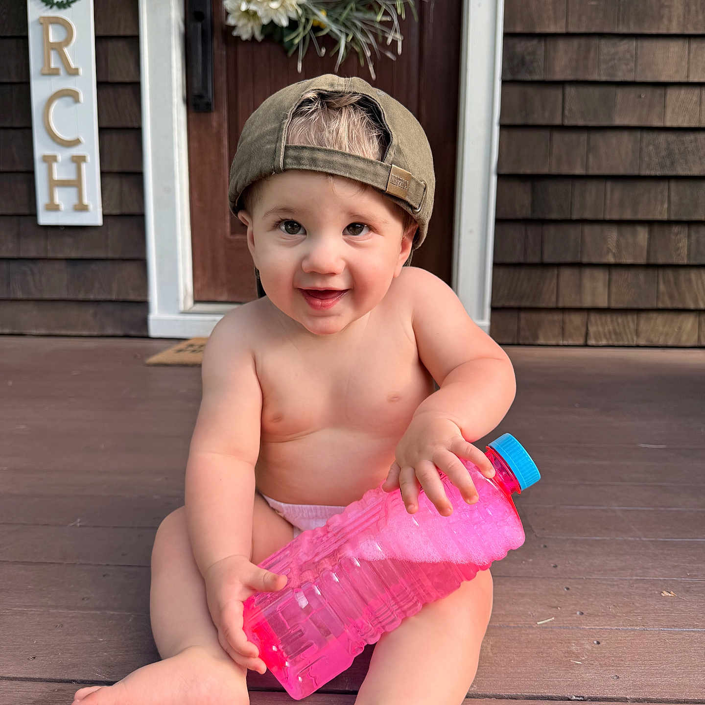 Alexander is registered to the contest to win money with this photo: baby, child, smiling, cap, water_bottle, porch, wooden_floor, wreath, door, sign, happy, sitting, outdoor, cute, infant, bare_chest, playful, person, holding, summer