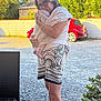 woman, baby, babywearing, outdoor, greenery, red_car, gravel, shorts, glasses, footwear, plant, sunlight, holding, embrace, headwear, barefoot, fabric, house, love, nature