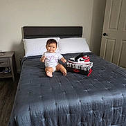 Samantha is registered to the contest to win money with this photo: baby, child, bed, comforter, sneaker, shoe_box, pillow, nightstand, door, floor, smiling, white_shirt, unicorn, indoor, headboard, footwear, person, home, clothing, furniture