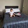 baby, child, bed, comforter, sneaker, shoe_box, pillow, nightstand, door, floor, smiling, white_shirt, unicorn, indoor, headboard, footwear, person, home, clothing, furniture