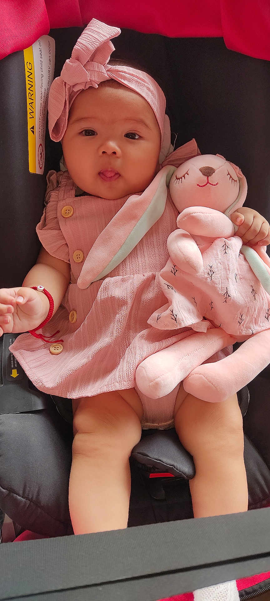 Samantha joined the competition — help win amazing prizes! baby, infant, car_seat, pink_dress, headband, plush_toy, bunny, toy, child, cute, playful, red_bracelet, warning_label, car_seat_strap, closeup, portrait, sitting, indoor, soft_toy, adorable