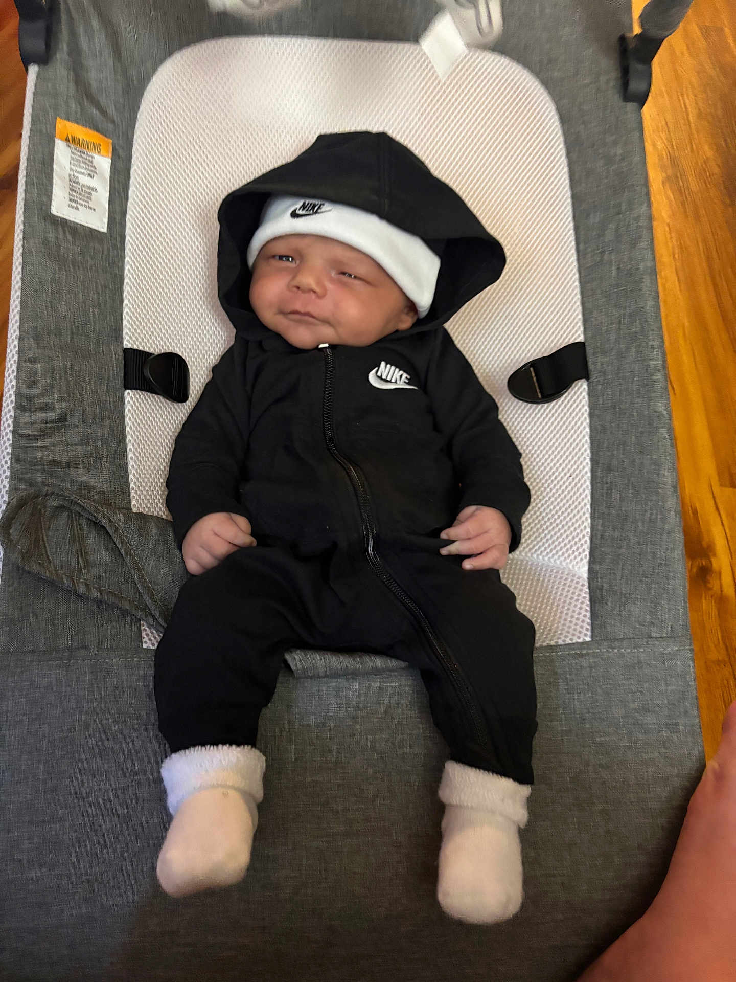 Kamarion is registered to the contest to win money with this photo: baby, infant, nike, hoodie, hat, socks, baby_rocker, gray, white, black_clothing, indoor, footwear, cute, child, resting, portrait, smile, person, apparel, relaxing