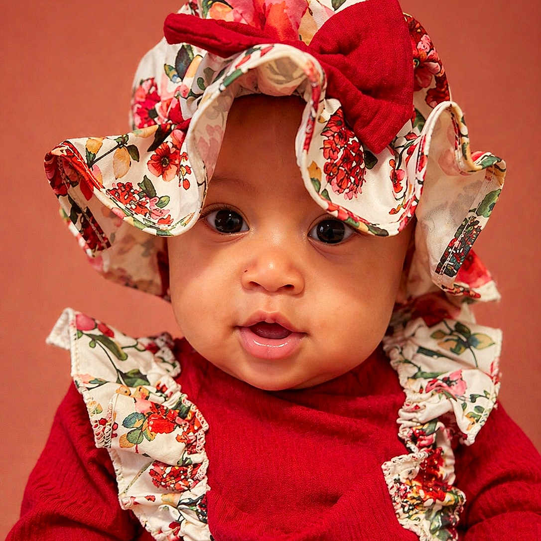 Navayla is registered to the contest to win money with this photo: adorable, baby, background, bow, child, closeup, cute, expression, eyes, face, fashion, floral, hat, headwear, infant, portrait, red_clothing, smile, warm_colors, young