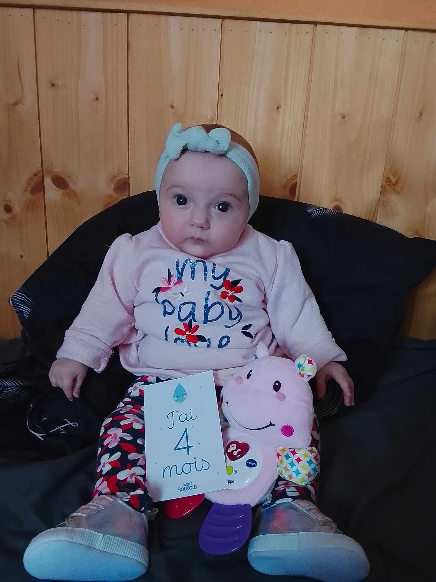 Manon a rejoint le concours — aidez-le/la à gagner de superbes lots ! baby, infant, headband, pink_clothing, toy, hippo_toy, card, wooden_wall, sitting, floor_cushion, shoes, floral_pants, indoor, child, cute, portrait, young_child, expression, colorful, clothing