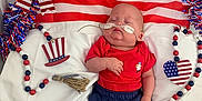 Asher is registered to the contest to win money with this photo: american_flag, baby, beads, blue_clothing, celebration, child, cute, hat_decoration, heart_shape, hospital_bed, indoor, infant, medical_equipment, nasal_cannula, patriotic_decor, person, red_clothing, sleeping, tassel, white_cushion