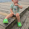 toddler, child, water_bottle, cap, green_clothing, green_slippers, dock, wooden_dock, water, boats, buildings, overcast_sky, outdoor, summer, sitting, curious_expression, sideways_cap, casual, daytime, portrait