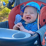 Dakota is registered to the contest to win money with this photo: baby, smiling, stroller, hat, blue_clothing, outdoor, sunlight, greenery, happy, child, face, hands, infant, seat, baby_toy, casual, daylight, cute, young_child, portrait
