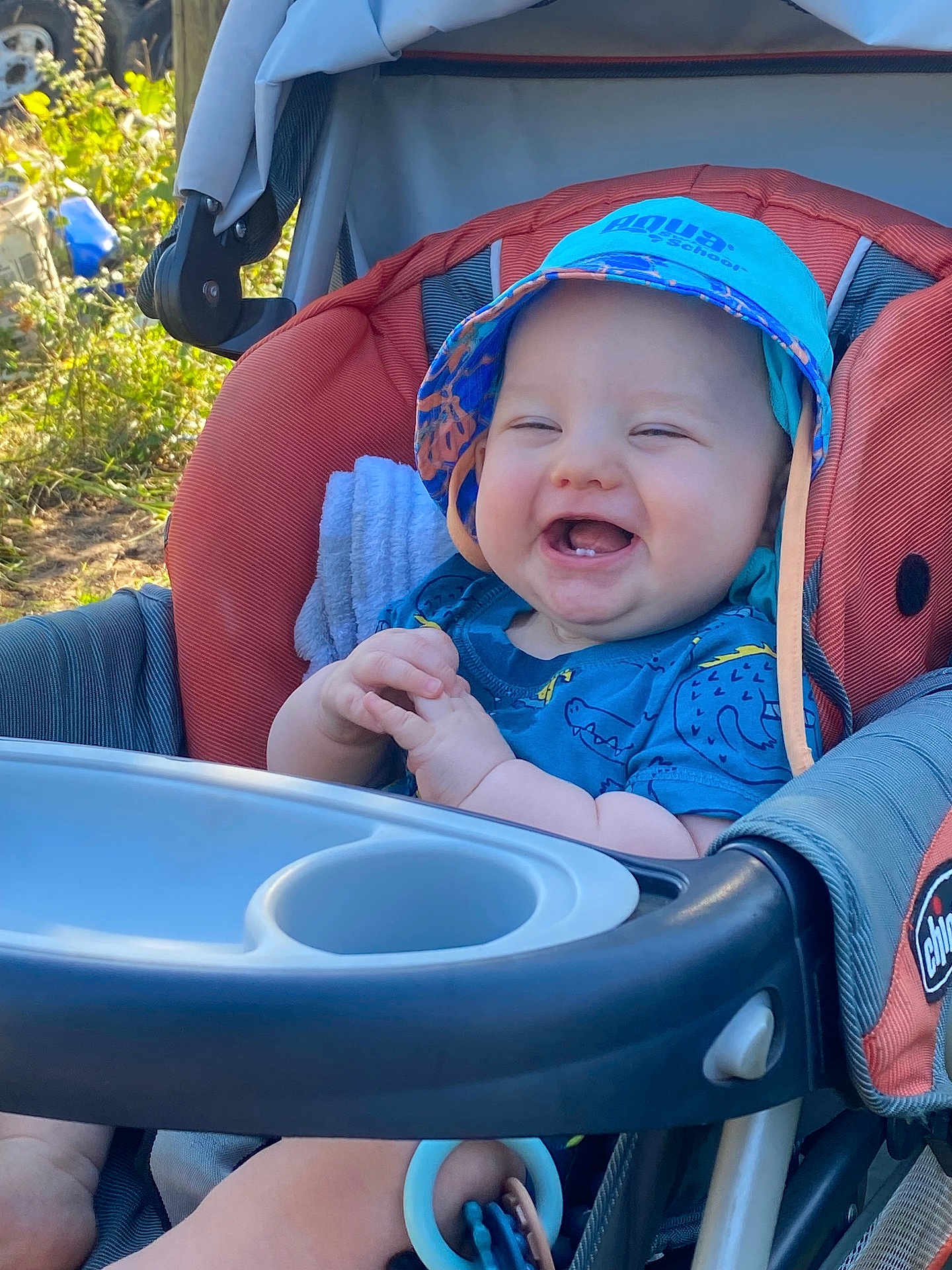 Dakota is registered to the contest to win money with this photo: baby, smiling, stroller, hat, blue_clothing, outdoor, sunlight, greenery, happy, child, face, hands, infant, seat, baby_toy, casual, daylight, cute, young_child, portrait