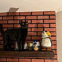 black_cat, cat, brick_wall, wooden_shelf, star_wars_figures, figurines, shadow, yellow_eyes, indoor, pet, animal, curious, toy, decor, shelf, wall, collectibles, standing, feline, wide_eyes