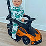 toddler, child, toy_car, indoor, floor, smiling, dinosaur_pajamas, handle, footwear, play, vehicle, black_handle, orange_toy, happy, young_child, flooring, casual, baby, transport, ride_on_toy