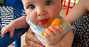 Carter is registered to the contest to win money with this photo: baby, child, cheese_puff, food, hand, face, mouth, outdoor, table, chair, adult_hand, clothing, skin, eyes, expression, person, candid, summer, snack, cute