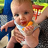 baby, child, cheese_puff, food, hand, face, mouth, outdoor, table, chair, adult_hand, clothing, skin, eyes, expression, person, candid, summer, snack, cute