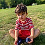 child, boy, grass, outdoor, sitting, red_shorts, striped_shirt, socks, cute, sunlight, nature, portrait, young, casual_clothing, daylight, playful, greenery, expression, knees_up, summer