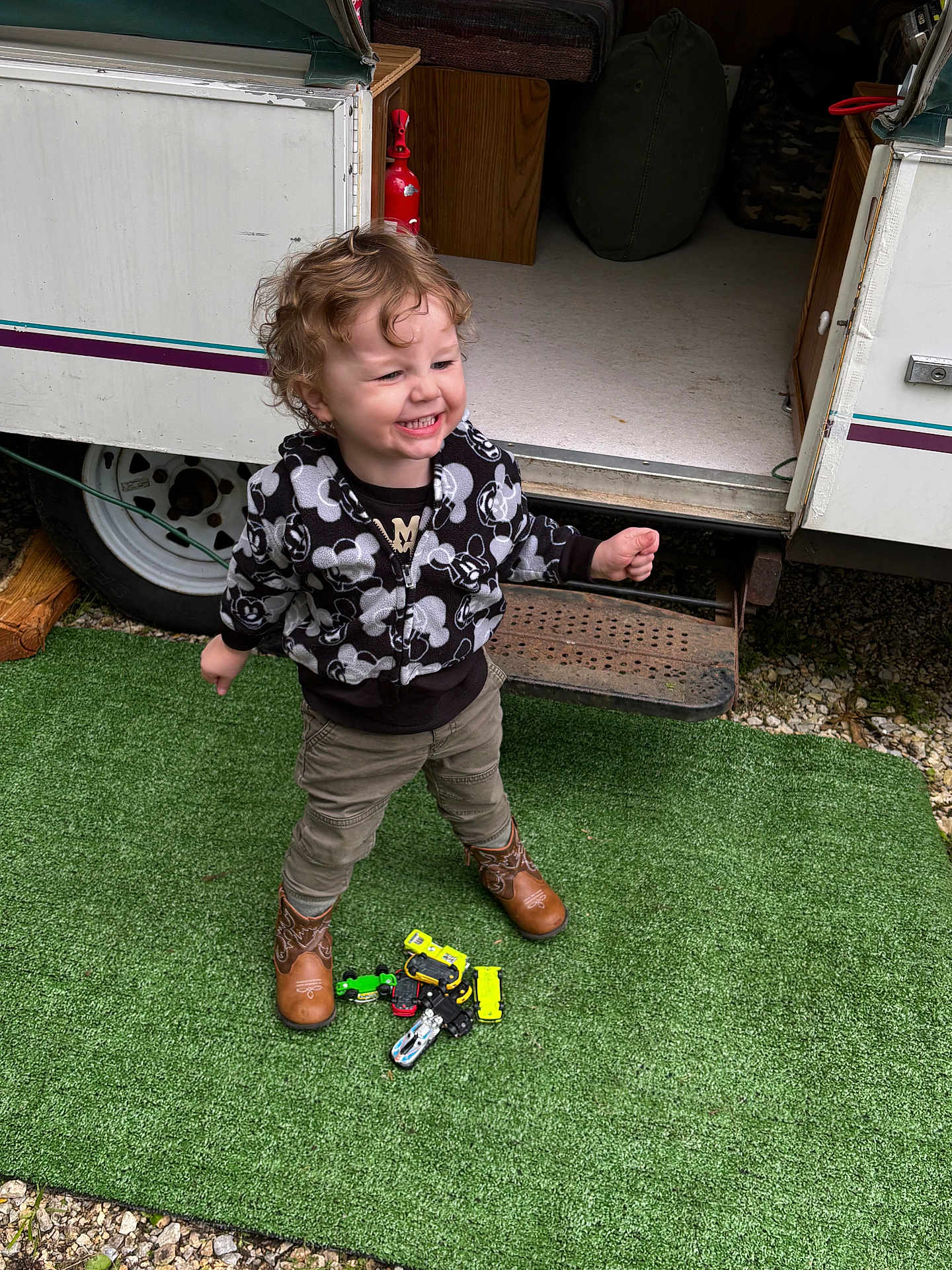 Kade is registered to the contest to win money with this photo: child, toddler, curly_hair, smiling, toy_cars, boots, jacket, outdoor, camper, green_mat, playing, happy, person, footwear, vehicle_toys, door, step, gravel, clothing, standing