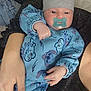 arm, baby, beanie, blue_onesie, cartoon_print, child, comfort, cuddle, cute, face, hand, headwear, indoor, infant, newborn, pacifier, person, portrait, sleepwear, sparkly_clothing