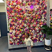 Annie is registered to the contest to win money with this photo: child, flower_wall, indoor, plant, neon_sign, colorful, cozy_clothing, curious, floor, door, potted_plant, leggings, jacket, sneakers, decor, artificial_flowers, wall, standing, portrait, casual