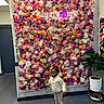 child, flower_wall, indoor, plant, neon_sign, colorful, cozy_clothing, curious, floor, door, potted_plant, leggings, jacket, sneakers, decor, artificial_flowers, wall, standing, portrait, casual