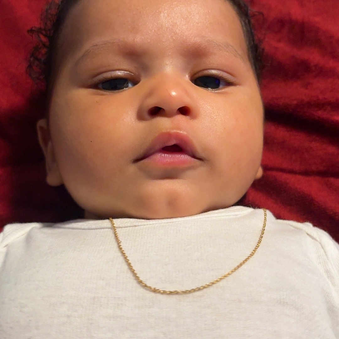 Akhyis is registered to the contest to win money with this photo: baby, closeup, curly_hair, cute, expression, face, gold_chain, head, indoors, infant, looking_at_camera, lying_down, newborn, peaceful, person, portrait, red_blanket, skin, white_clothing, young_child